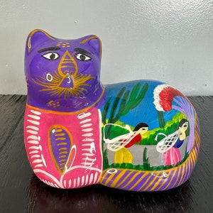 May include: A hand-painted ceramic cat figurine with a purple, pink, and yellow design. The cat is lying down with its head turned to the side. The figurine is decorated with a scene of people and cacti in a desert landscape.