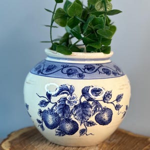 May include: A cream-colored ceramic vase with blue floral designs, including strawberries and leaves, sits on a wooden slice. The vase has a blue rim and is filled with green leafy stems, creating a decorative arrangement.
