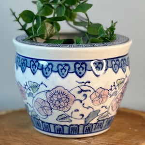 May include: A ceramic planter with a floral design in pink and blue, filled with green foliage. The planter sits on a wooden slice. The planter has a white base with blue floral patterns and a blue rim.