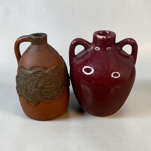 May include: Two ceramic vases. One is brown with a handle and a decorative band. The other is a deep red with two handles. Both have a rustic, antique appearance. The vases are on a plain, light background.