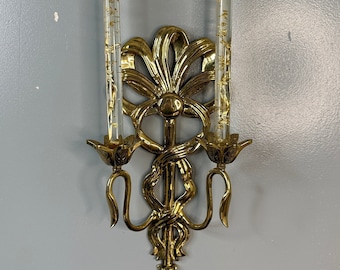 May include: A pair of gold-toned wall sconces with ornate designs and clear glass candle holders. The sconces feature a ribbon motif and are designed to hold two candles each.