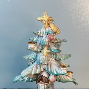May include: A ceramic Christmas tree figurine with a white base, green and blue branches, and gold star on top. The tree is decorated with small gold stars and red and pink ornaments.