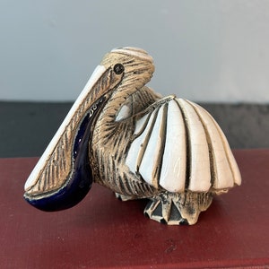 May include: A small ceramic pelican figurine with a white body, brown wings, and a blue beak. The figurine is sitting on a red book.