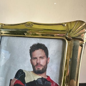 May include: A gold-colored picture frame with a decorative shell design. The frame holds a photo of a person wearing a colorful sweater and blue jeans.