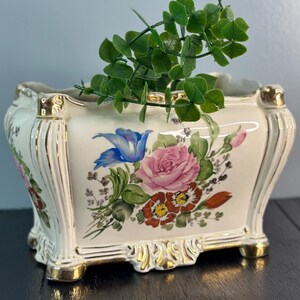 May include: A white ceramic planter with gold trim and floral designs. The planter has a square shape with rounded corners and four feet. It is decorated with a pink rose, blue flower, and other colorful flowers. A small green plant is inside the planter.