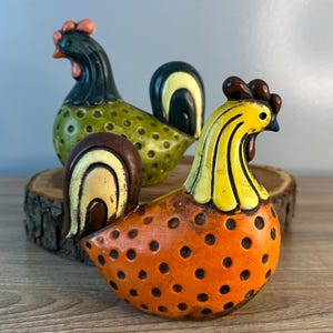 May include: Three ceramic rooster figurines with polka dot patterns. The rooster on the left is green with black accents, the rooster in the middle is orange with yellow accents, and the rooster on the right is yellow with brown accents.