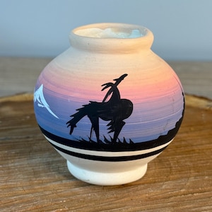 May include: A small, round ceramic vase with a painted landscape scene. The vase features a sunset gradient of pink, purple, and blue. A black silhouette of a horse and rider is painted on the side. The vase has a white base and a light tan rim.