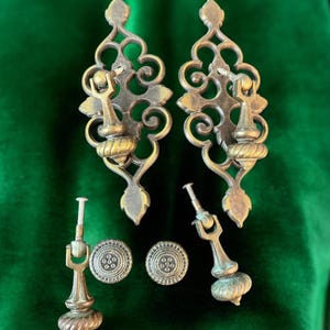 May include: A collection of antique brass hardware, including two ornate pulls with scrolling designs and two smaller pulls with decorative knobs. Also included are two round, patterned cabinet knobs. The hardware is displayed on a green velvet surface.
