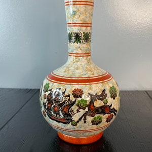 May include: A white ceramic vase with a geometric design and a deer and ship scene. The vase has a speckled glaze and orange accents.