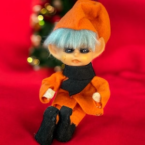 May include: A vintage doll with an orange felt hat and outfit, blue hair, and black boots. The doll has a black scarf and is sitting on a red surface. A small Christmas tree is in the background.