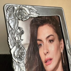 May include: A silver picture frame with a detailed rose design. The frame is holding a photo of a woman with long dark hair. The woman is wearing a silver necklace and earrings.