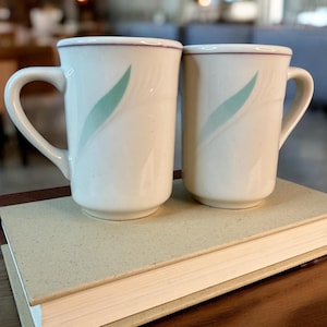 May include: Two white ceramic mugs with a teal leaf design and a purple rim, resting on a light brown book. The mugs have curved handles and are set against a blurred background.