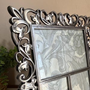 May include: A decorative silver-colored picture frame with an ornate floral design. The frame holds three sections, each displaying a light gray floral pattern. The frame is made of metal and has a detailed, textured surface.
