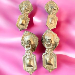 May include: Four antique brass drawer pulls with ornate geometric designs. Each pull features a rectangular base and a decorative top with a loop for attaching to a drawer. The pulls are arranged on a pink background.