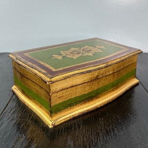 May include: A small wooden box with a gold and green painted design. The box has a curved edge and a decorative floral pattern on the top.