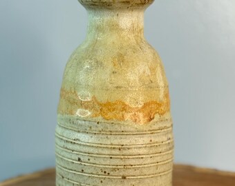 Puede incluir: Un jarrón de cerámica hecho a mano con una superficie texturizada y un cuello estrecho. El jarrón presenta ranuras horizontales y una paleta de colores beige, marrón claro y marrón. El jarrón se exhibe sobre una rodaja de madera.