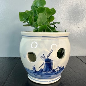 May include: A white ceramic planter with a blue and white windmill design. The planter has drainage holes and is filled with green plants.