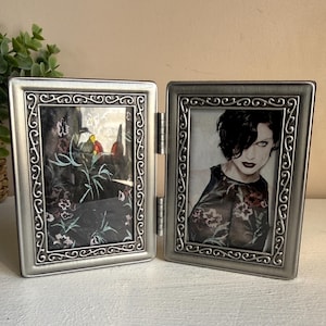 May include: A hinged, silver-colored double picture frame. Each frame has a decorative scroll design around the edges. One frame holds a picture of a person with dark hair and red lipstick. The other frame holds a picture of a floral design.