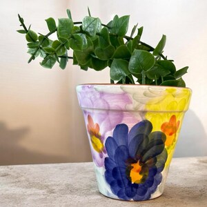 May include: A hand-painted ceramic flower pot with a vibrant floral design. The pot features a large blue flower, yellow and orange accents, and a green plant. The pot is filled with a green leafy plant, adding a touch of nature.