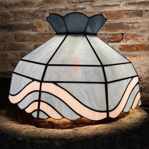 May include: A stained glass lamp shade with a light blue and white design. The shade has a scalloped edge and a geometric pattern with black lines. The lamp sits on a stone surface against a brick wall.