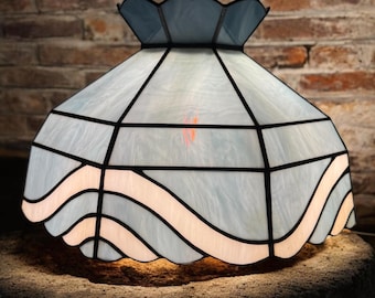 Puede incluir: Una pantalla de lámpara de vidrieras con un diseño azul claro y blanco. La pantalla tiene un borde festoneado y un patrón geométrico con líneas negras. La lámpara está sobre una superficie de piedra contra una pared de ladrillo.