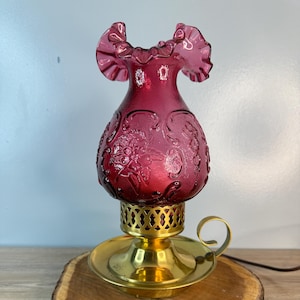 May include: A vintage ruby red glass lamp with a ruffled top and floral embossed design. The lamp has a brass base and sits on a wooden slice. The lamp is a decorative lighting piece.