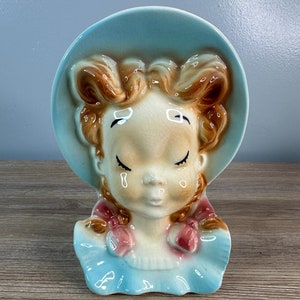 May include: A vintage ceramic planter shaped like a girl's head with closed eyes, wearing a blue hat and a pink bow. The planter is made of glazed ceramic and has a light blue and white color scheme.