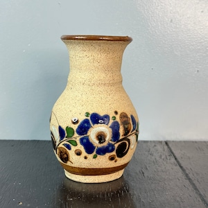 May include: A small, cream-colored ceramic vase with a brown rim and a floral design in shades of blue, green, and brown.
