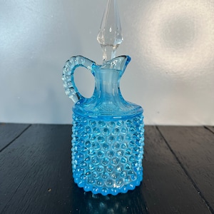 May include: A light blue glass bottle with a textured, bubbled surface and a clear glass stopper. The bottle has a handle and a rounded base.