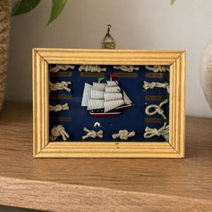 May include: A framed nautical display featuring a miniature sailboat with white sails and a red hull, set against a dark blue background. The frame is gold-colored, and the display includes various rope knots. A decorative piece.