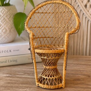May include: A miniature wicker chair with a rounded back and a woven seat. The chair is made of light brown wicker and has a decorative black and white trim. The chair is placed on a wooden surface.