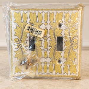 May include: A double light switch wall plate featuring a yellow and white acanthus leaf pattern. The rectangular plate has two vertical toggle switch openings and four screw holes. It is sealed in clear plastic packaging with a barcode sticker.