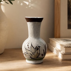 May include: A small, off-white ceramic vase with a bulbous body and a flared neck. The rim is painted in a dark brown tone. The front of the vase features a hand-painted illustration of a bird in black and green ink, set against a textured surface.