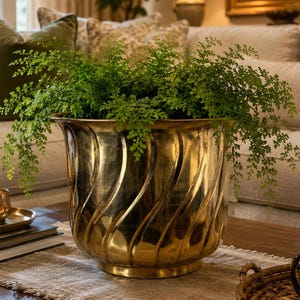 May include: A large, gold-colored planter with a swirling design holds a lush green fern. The planter sits on a wooden coffee table with a neutral-toned runner. The room has a warm, inviting atmosphere.