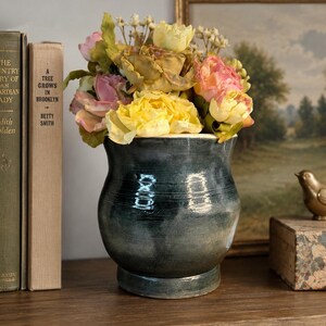 May include: A dark teal ceramic vase with a glossy finish and a rounded body, filled with a bouquet of artificial flowers in shades of yellow, pink, and cream. The vase features a wide opening and a sturdy base, showcasing subtle horizontal texture.