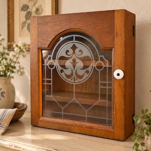 May include: A rectangular wooden wall cabinet with a hinged door featuring an arched glass panel. The glass is etched with a central circular floral motif and geometric lines. A small white ceramic knob with a black center is attached to the door.
