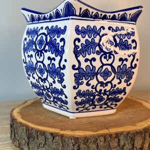 Puede incluir: Una maceta de cer&aacute;mica blanca con un estampado floral azul. La maceta tiene un borde festoneado &uacute;nico y una forma geom&eacute;trica. El dise&ntilde;o azul y blanco recuerda a la porcelana cl&aacute;sica. La maceta est&aacute; sobre una rodaja de madera.