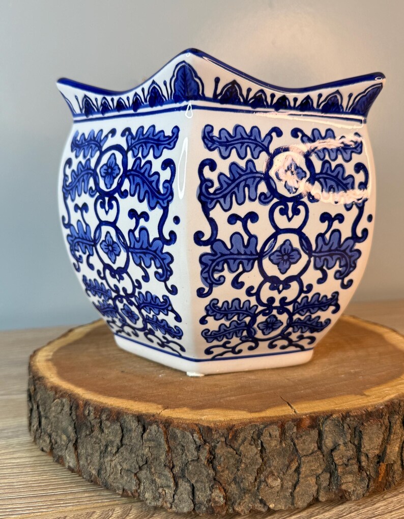 Puede incluir: Una maceta de cer&aacute;mica blanca con un estampado floral azul. La maceta tiene un borde festoneado &uacute;nico y una forma geom&eacute;trica. El dise&ntilde;o azul y blanco recuerda a la porcelana cl&aacute;sica. La maceta est&aacute; sobre una rodaja de madera.