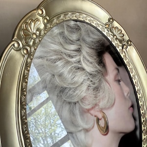 May include: An oval-shaped, gold-colored picture frame with ornate detailing. The frame surrounds a portrait of a person with blonde hair and gold hoop earrings. The background shows a reflection of a tree and sky.