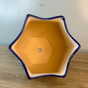 Puede incluir: Una maceta de cer&aacute;mica en forma de estrella con borde azul e interior blanco. La maceta tiene un color terracota natural en el interior y un orificio de drenaje en la parte inferior. Dise&ntilde;ada para uso en interiores o exteriores.
