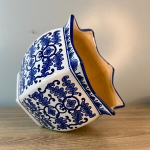 Puede incluir: Una maceta de cer&aacute;mica blanca con un dise&ntilde;o floral azul. La maceta tiene un borde festoneado &uacute;nico y un interior marr&oacute;n claro. El patr&oacute;n azul es intrincado y cubre el exterior de la maceta, creando una pieza decorativa.