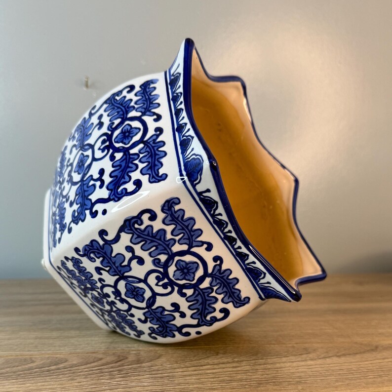 Puede incluir: Una maceta de cer&aacute;mica blanca con un dise&ntilde;o floral azul. La maceta tiene un borde festoneado &uacute;nico y un interior marr&oacute;n claro. El patr&oacute;n azul es intrincado y cubre el exterior de la maceta, creando una pieza decorativa.