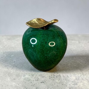May include: A decorative apple sculpture with a polished, deep green body and a gold-colored leaf. The apple has a smooth, rounded shape, and the leaf is detailed with veining. The sculpture is a unique home decor item.