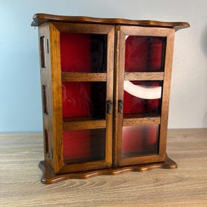 May include: A small, wooden curio cabinet with two glass doors. The cabinet has a dark brown finish and a red velvet interior. The doors have small, ornate handles. The cabinet is sitting on a wooden surface.