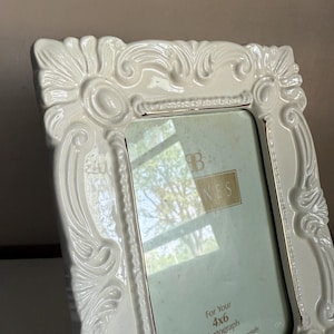 May include: A white, ornate picture frame with a glossy finish. The frame has a detailed, embossed design and is sized for a 4x6 photograph. The frame is designed to display the photo vertically or horizontally.