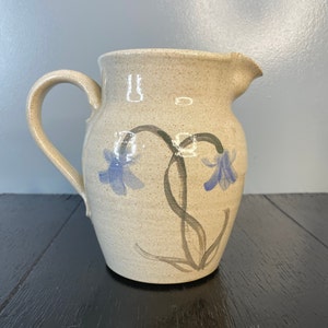 May include: A beige ceramic pitcher with a handle and spout. The pitcher is decorated with blue flower illustrations and gray stems. The pitcher is sitting on a black surface.