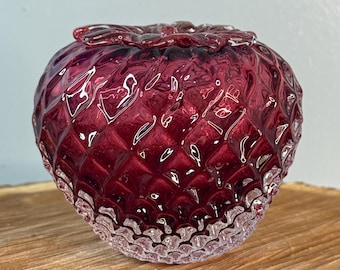 Puede incluir: Un tarro de vidrio redondo, rojo rubí, con un cuerpo texturizado con forma de diamante y una base transparente y festoneada. El tarro tiene una tapa a juego con un diseño floral. El tarro mide aproximadamente 10 cm de diámetro.