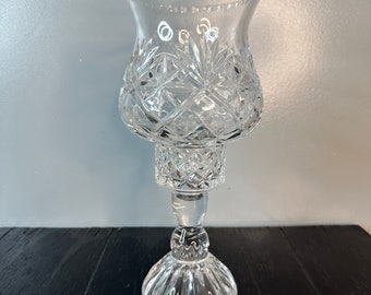May include: A clear glass candle holder with a cut-glass design. The holder has a wide base and a tall, slender stem.