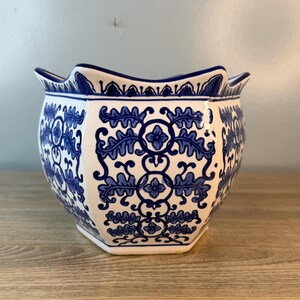 Puede incluir: Una maceta de cer&aacute;mica blanca con un estampado floral azul. La maceta hexagonal tiene un borde festoneado y est&aacute; decorada con intrincados dise&ntilde;os de hojas y flores azules. La maceta est&aacute; sobre una superficie de madera.