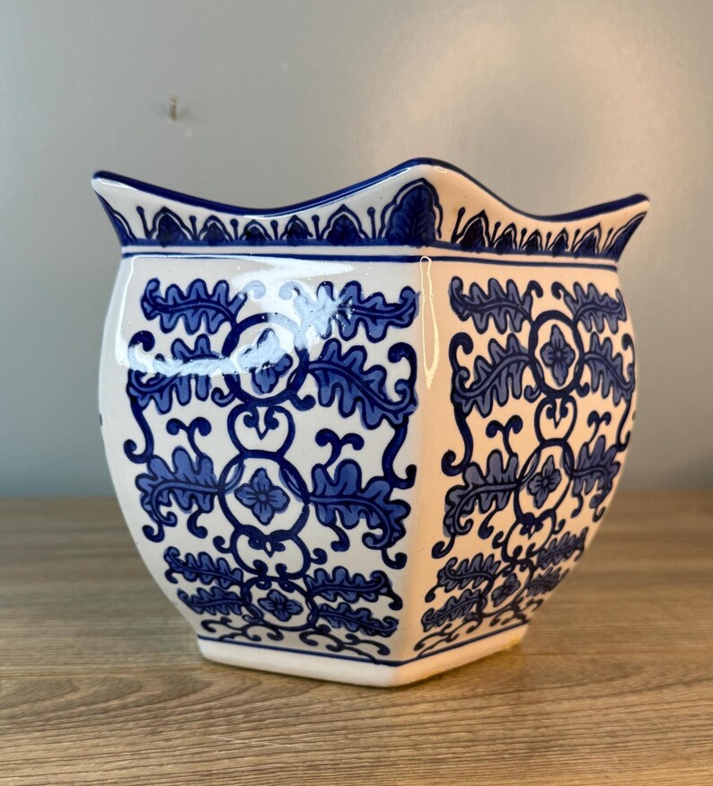 Puede incluir: Una maceta de cer&aacute;mica blanca con un estampado floral azul. La maceta tiene un borde festoneado y una forma geom&eacute;trica. El dise&ntilde;o azul presenta hojas y motivos florales. Adecuado para uso en interiores o exteriores.